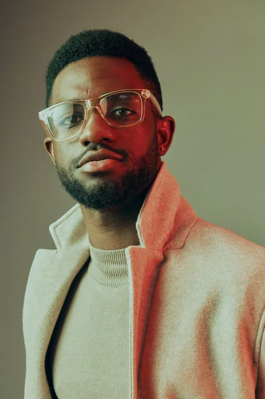 Portrait of a stylish Black man wearing glasses and a light coat, exuding confidence with a warm smile against a muted background, perfect for lifestyle and fashion themes