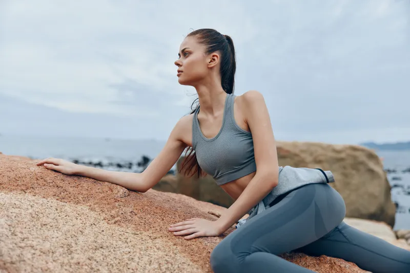 Young athletic woman in activewear poses on a rocky coastline, showcasing strength and tranquility against a grey ocean backdrop during a peaceful moment