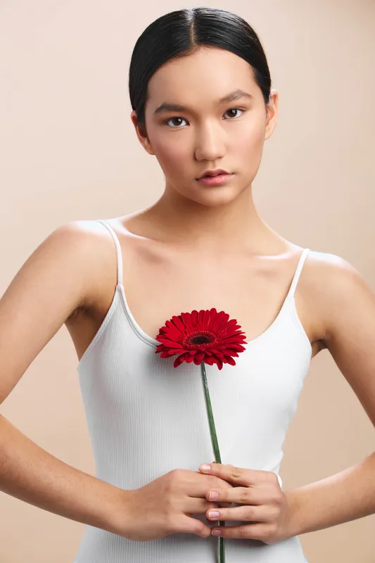Asian woman holds a red flower against a soft beige backdrop, wearing a white tank. Her calm expression conveys elegance, confidence, and a minimalist, timeless beauty.