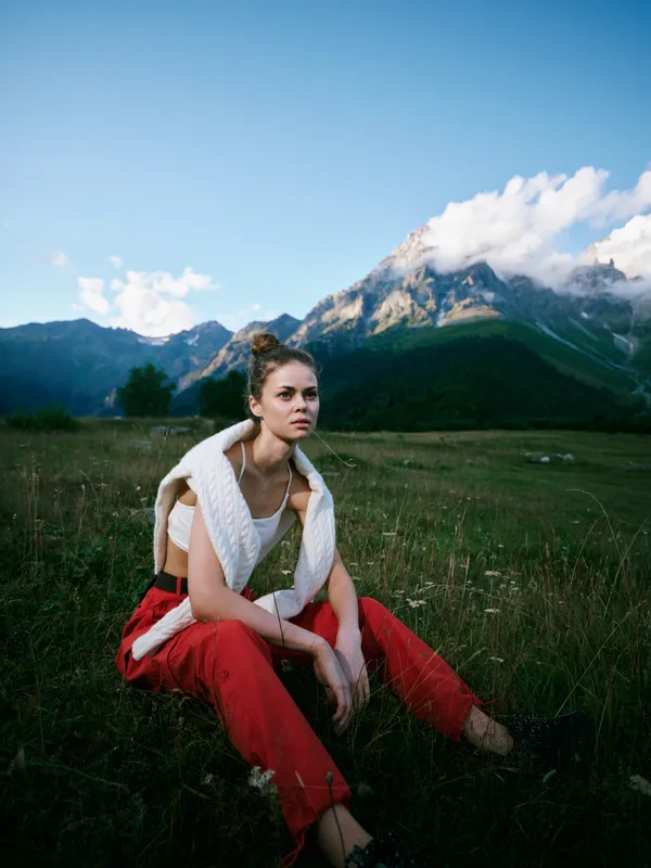 Outdoor scene with a woman seated in a meadow, mountains in the distance, red pants and knit scarf, casual fashion and tranquil natural landscape