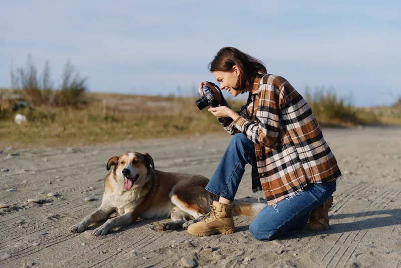 Photographer kneels on a sandy beach, dog beside, capturing a candid outdoor moment in a wide open landscape under a clear sky with soft light and joyful connection.