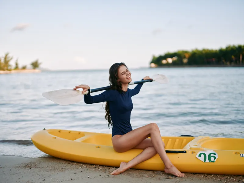 Female surfer sits on a bright yellow kayak at the shoreline, stretching arms with a carefree smile as calm water and distant trees frame a sunny beach scene.