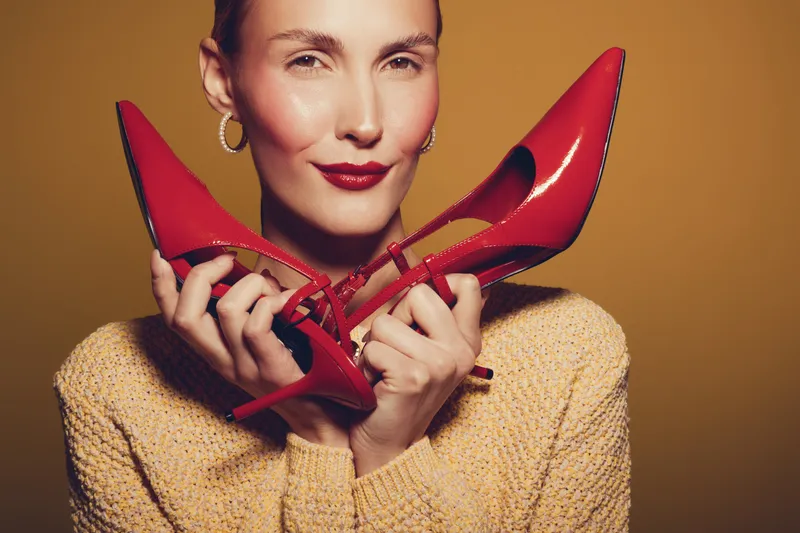Woman in yellow sweater holds red high heels near face, smiling softly. Studio portrait with warm lighting on brown background. Concept of fashion, style, and elegance.
