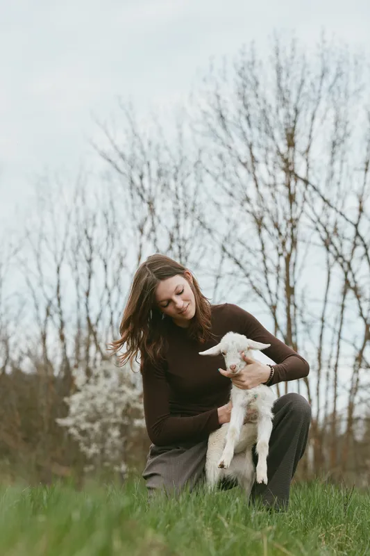 A woman sits in a grassy field, calmly holding a small goat kid, smiling softly. Natural light, serene mood, highlighting care, companionship, and everyday animal affection.