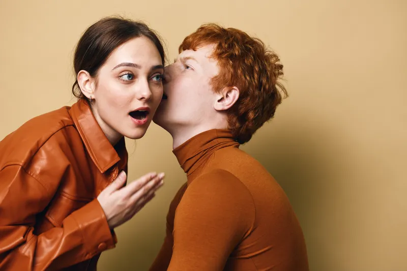 Man whispering secret to surprised woman in brown clothing on beige background. Emotional expression, casual outfit, communication between young adults in intimate moment.
