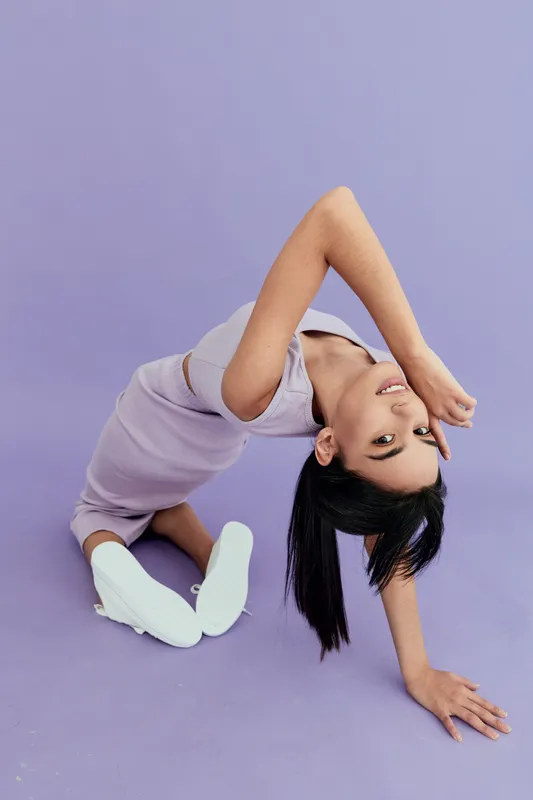 flexible young woman in lavender outfit posing on violet background, wearing white sneakers and showing modern dance or yoga stretch.