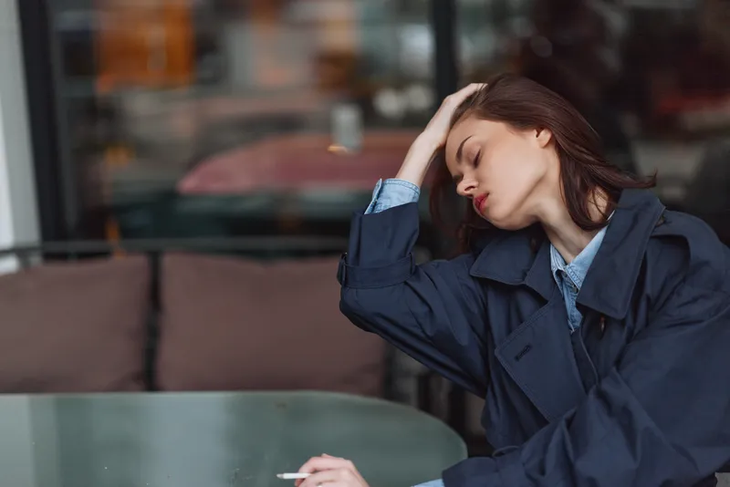 A tired woman rests her head on her hand at an outdoor cafe, denim jacket draped over her shoulders, a cigarette on the table creating a moody, introspective scene.