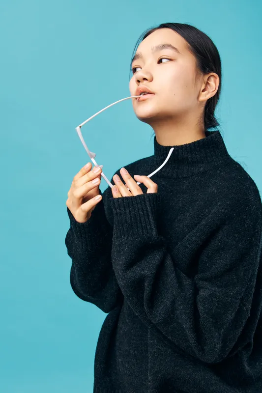 Young woman wearing black sweater holding glasses near face with thoughtful expression. Portrait with blue background, casual fashion and minimalistic style for lifestyle concept.