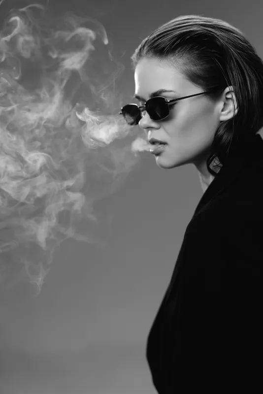Black and white portrait of a stylish woman with sunglasses exhaling smoke, dressed in dark clothing, showing calm and intensity.