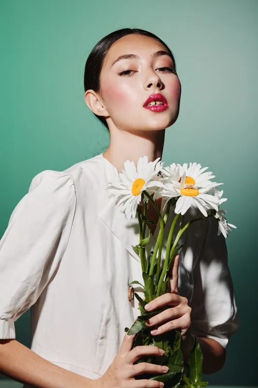 Young stylish woman with vibrant red lipstick holding a bouquet of white daisies. She poses confidently against a mint green background. Concept of beauty, fashion, and floral elegance.