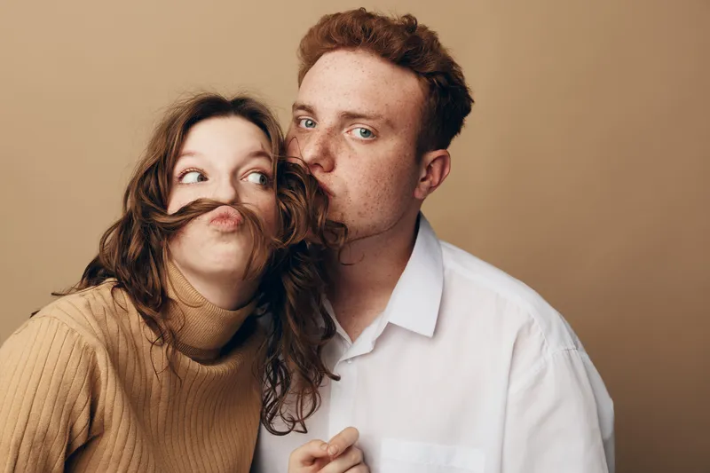 Playful young couple with curly hair and red hair posing together on beige background. Woman makes funny face with hair mustache while man kisses her cheek. Concept of love and fun.