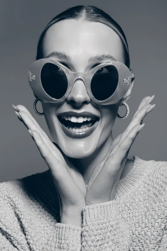 Young Caucasian woman with pale skin and slicked back hair wears round sunglasses and smiles widely, hands framing her face in a joyful expression.