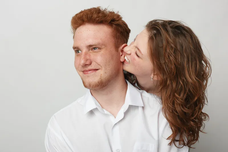 Happy young couple in white shirts share a playful moment, woman whispering in man’s ear. Studio portrait on light background, concept of love, friendship, and joyful relationships.