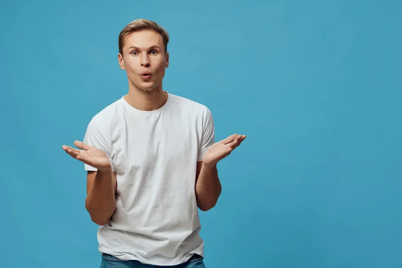 Confused young man with puzzled expression, wearing casual white t-shirt, gesturing with palms up, standing against bright blue background, studio shot. People lifestyle concept