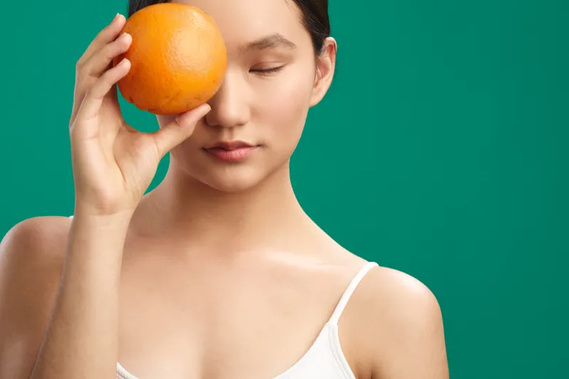 asian woman holds an orange near her eye in a vibrant studio, smiling softly as she explores color contrast and balance, creating a playful yet calm lifestyle portrait.