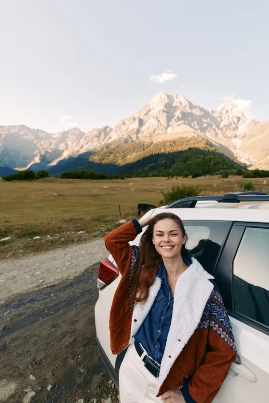 Woman smiling and leaning on white car by mountain road, travel portrait with scenic landscape backdrop. Casual jacket, outdoor adventure and happy road trip moment.