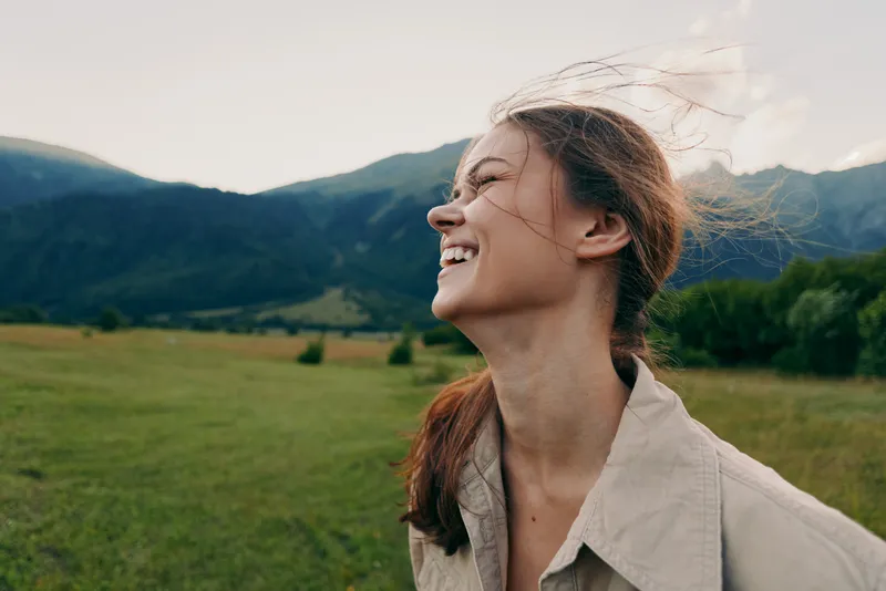 Young woman with long brunette hair laughs in the breeze in an open field with mountains in the background, her casual beige jacket catching the wind.