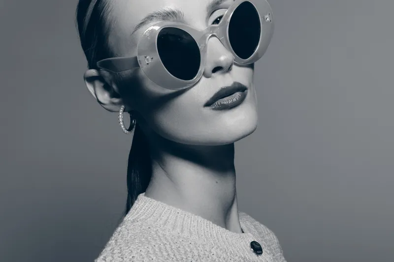 Portrait of a stylish Caucasian woman with pale skin wearing large round sunglasses and hoop earrings. She poses confidently in a textured sweater in studio lighting.