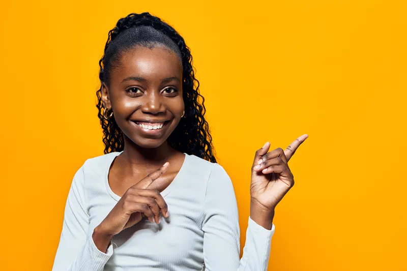 Happy young Black girl in casual attire pointing with a smile against a vibrant yellow background, promoting positivity and joy