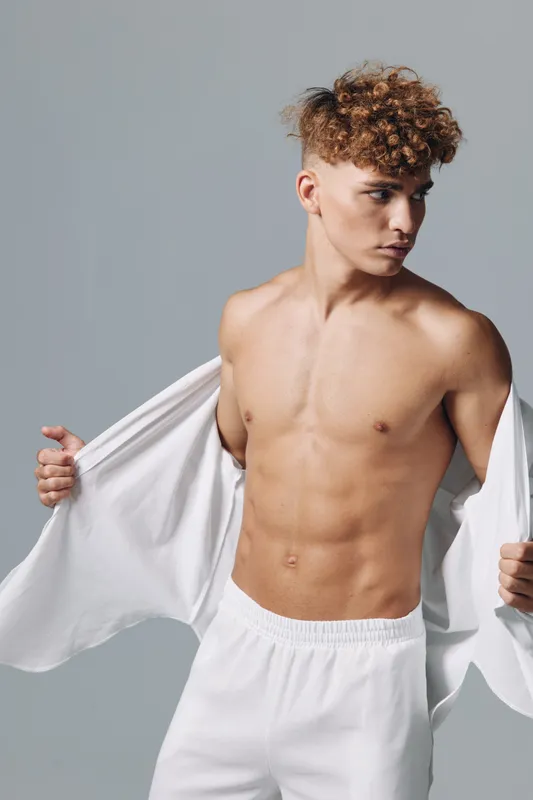 Young man with curly hair and muscular body taking off white shirt, posing in studio with neutral background, fitness and casual style concept.