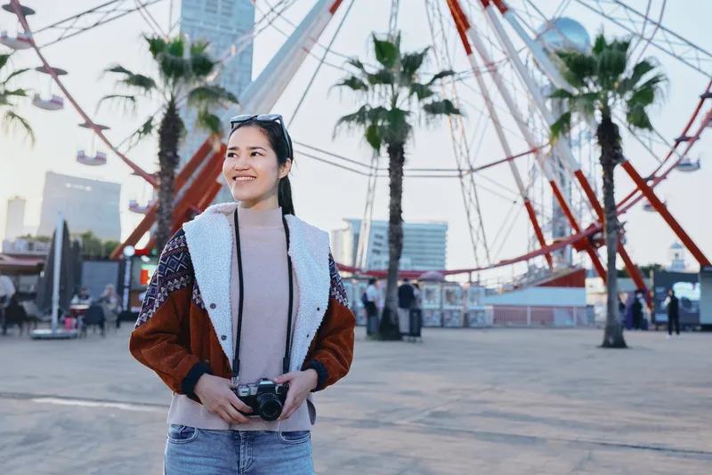 Camera in hand, a smiling woman stands beside a ferris wheel in a sunny urban plaza, city skyline behind, casual jacket and jeans, candid travel moment, leisure and street photography vibe for social