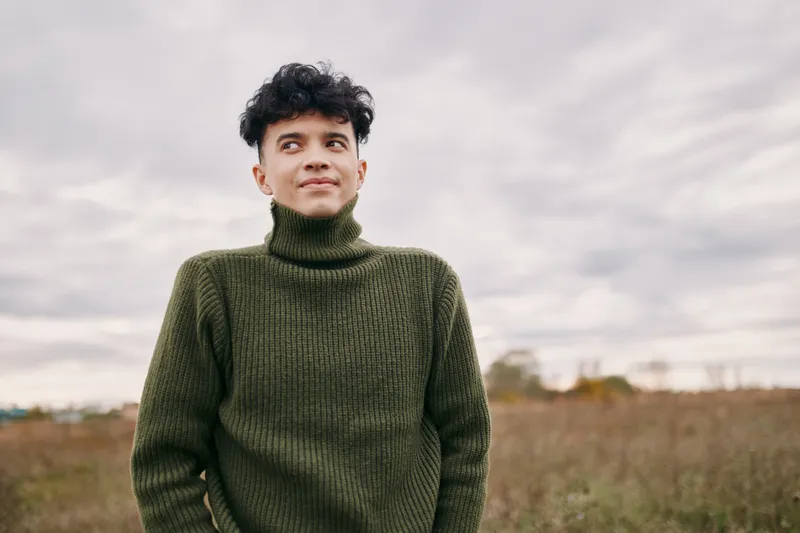 Young man wearing a green knit turtleneck stands in an open field, looking thoughtful and confident; authentic and genuine portrait capturing natural expression and relaxed posture.