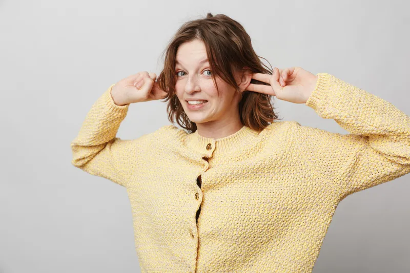 Young woman in yellow knitted sweater makes playful gesture with fingers near ears, smiling and looking at camera. Studio portrait on light grey background with casual style and fun mood.
