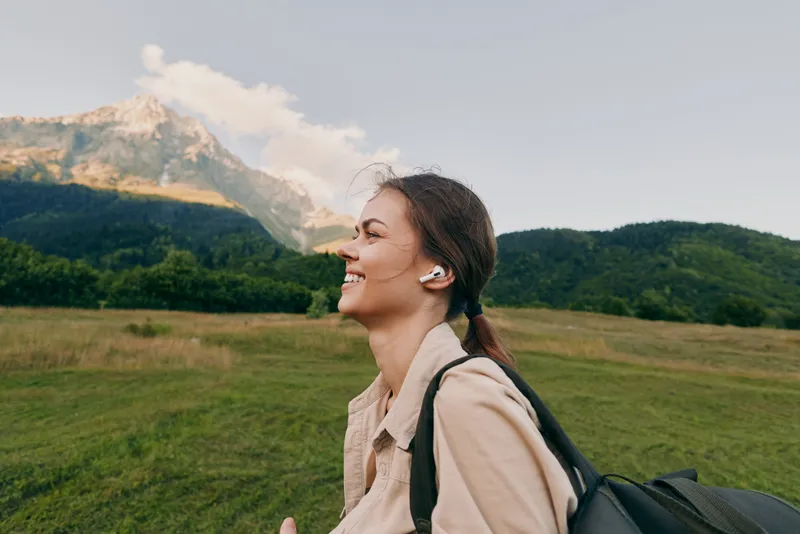 Smiling woman with backpack enjoying an outdoor stroll listening to earbuds. Expansive field and distant mountains, lifestyle and wellness vibe, natural daylight.