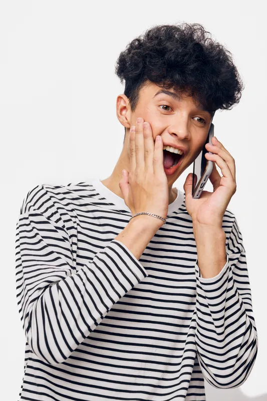 surprised young man talking on smartphone with striped shirt shows expression of shock and excitement against white background in casual style