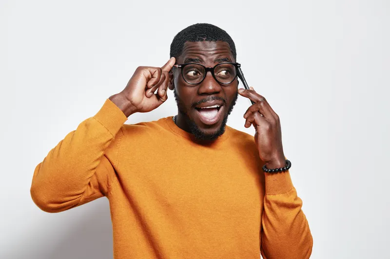 African American man with dark skin wearing glasses and orange sweater talking on smartphone with surprised expression isolated on white background. Communication and technology concept.