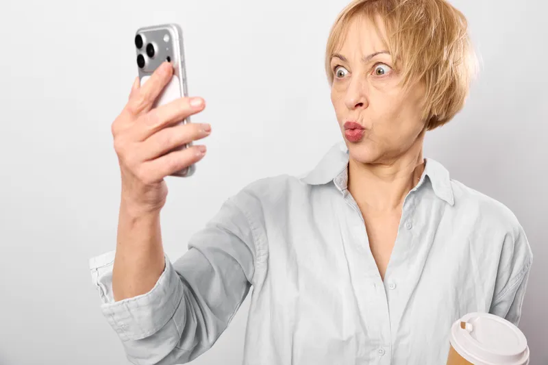 Older woman selfie with smartphone, surprised expression, holding coffee mug, studio portrait on white background, phone near face, casual grey shirt