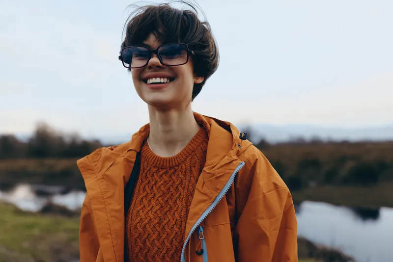 Smiling woman lifestyle portrait outdoors by a river, wearing orange jacket and glasses, holding a camera strap, soft morning light and calm nature background, fresh air breeze