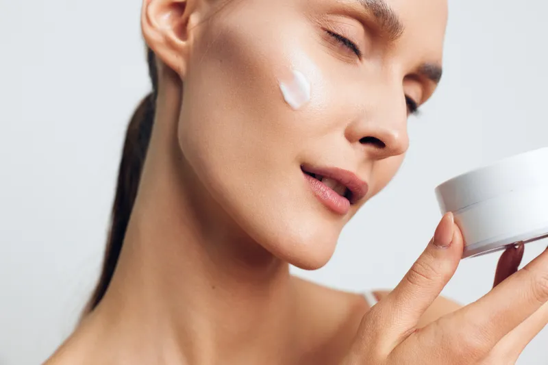 A young woman applies moisturizer to her cheek in a close-up, calm self-care moment, highlighting skincare routine and focused, gentle concentration.