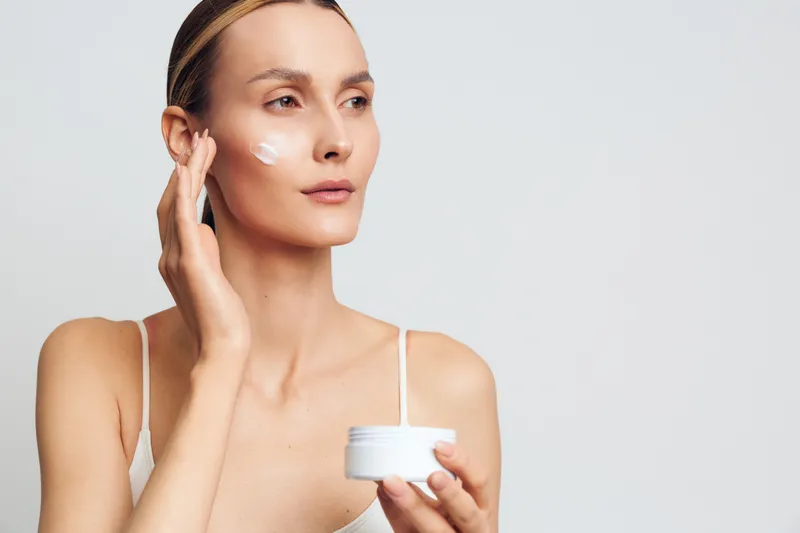 Young woman applying cream on face, skincare routine, natural beauty, clean skin, white background, soft lighting, focused expression, minimal makeup, health care concept