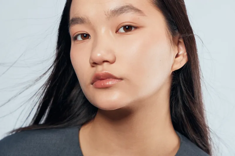 Portrait of young Asian woman with natural makeup and long dark hair wearing gray shirt against light background with serious expression and smooth clear skin in studio lighting.