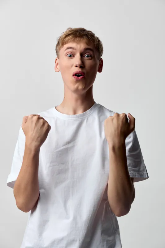Excited youth with fists raised in celebration, energetic expression and triumphant mood in a studio setting, white shirt, dynamic gesture, joyful anticipation for success