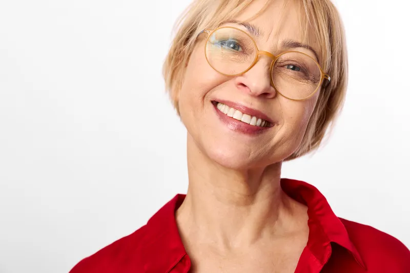 Smiling elderly woman portrait, closeup headshot with short blonde hair, wearing red shirt and eyeglasses, friendly expression, studio lighting, plain white background