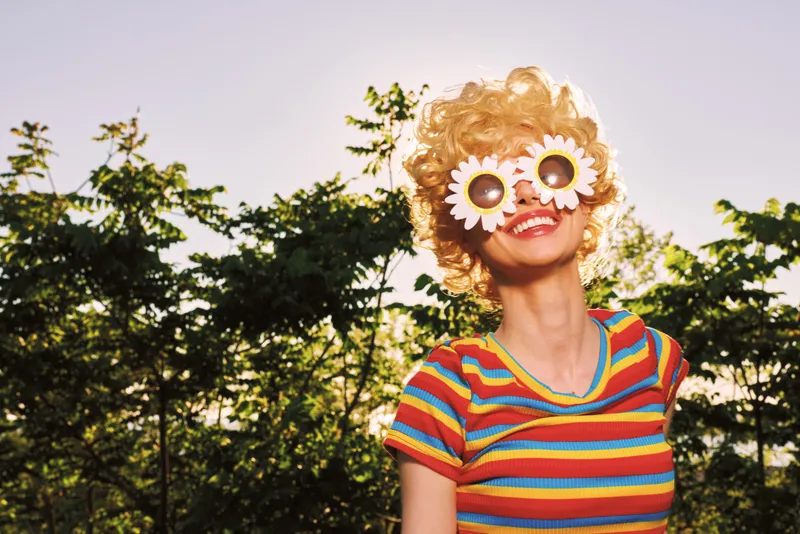 Cheerful woman outdoors, wearing a striped shirt and playful flower eye props, capturing a sunny, retro mood with vibrant colors and carefree laughter