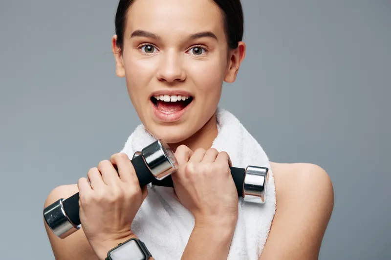 Fitness woman lifting dumbbells with a towel around her neck, showcasing strength and energy during a workout session in a clean gym setting, focused on health and active lifestyle for marketing.