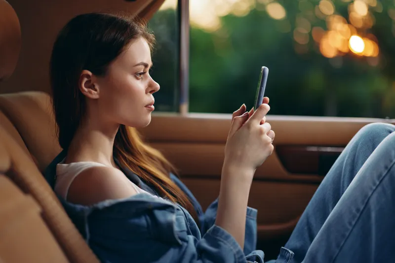 Young woman using smartphone in the car during sunset, wearing casual denim attire, with relaxed expression and nature background, conveying calmness and leisure.