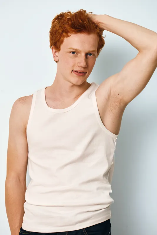Young man with red hair in white tank top posing indoors touching head with arm raised and looking at camera with slight smile on light background.