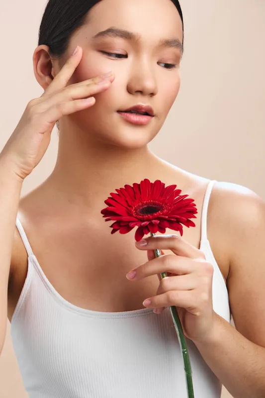 asian woman gently holds a vivid red flower, her calm gaze and soft smile create a serene portrait in a minimalist setting, highlighting beauty and quiet elegance.