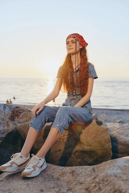 Lifestyle woman in modern boho-western denim street style sits on rocks at sunset by the sea, featuring film color tones and casual sneakers for relaxed outdoor vibe.