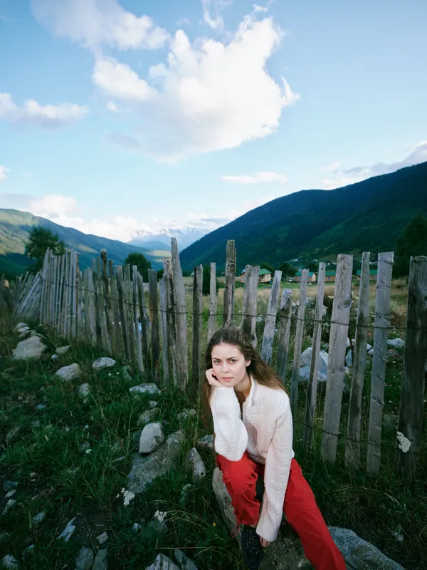 Woman posing by a rustic wooden fence in a mountainous landscape, casual outfit, red pants and white sweater, rocky terrain under a cloudy sky, serene outdoor setting for nature and fashion