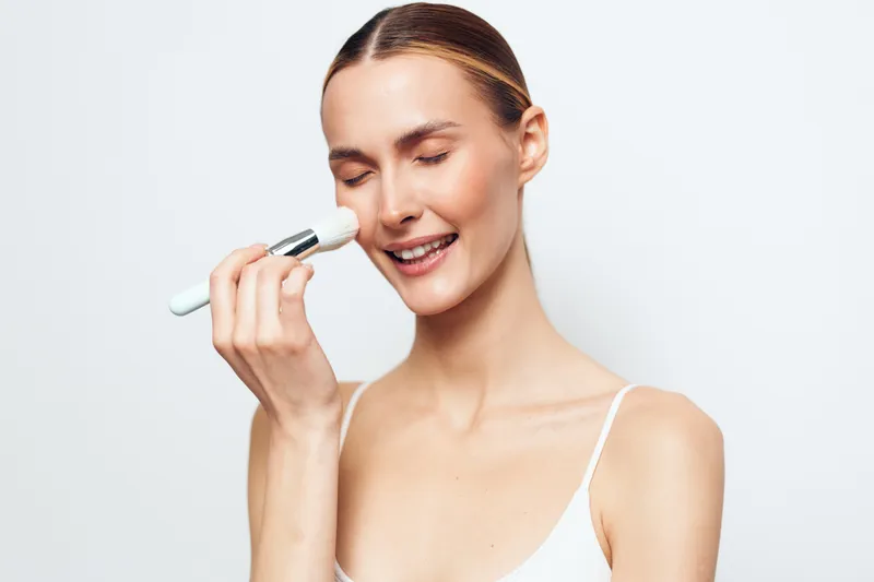 A smiling young woman with clear skin applies makeup powder on her face with a soft brush against a plain white background. Concept for beauty, skincare, and self-care routine.