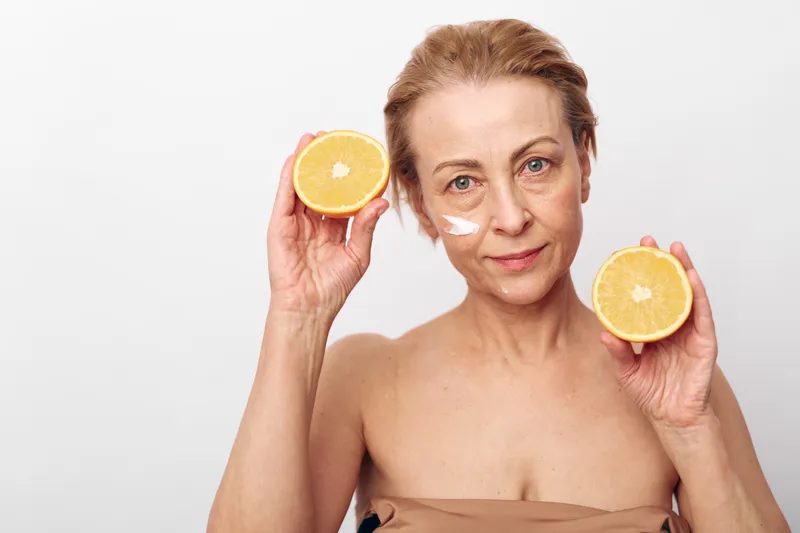 Mature woman with natural skin holds two halves of a fresh orange, applying cream on her face. Concept for skincare, healthy lifestyle, vitamin c benefits, and natural beauty care.