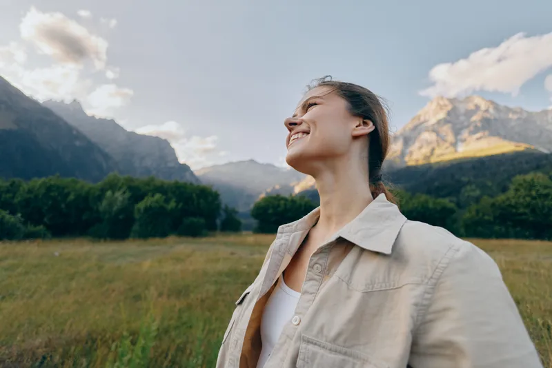 A smiling woman in a beige jacket stands in a grassy valley beneath towering mountains, enjoying fresh air and open space in a relaxed outdoor moment.