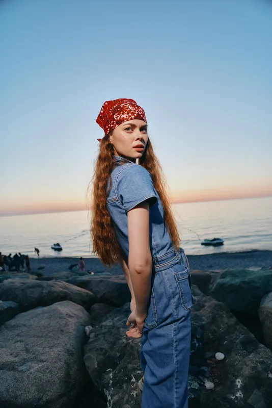 lifestyle woman in modern boho-western denim street style with red bandana by the beach at sunset in film color, relaxed and stylish outdoors