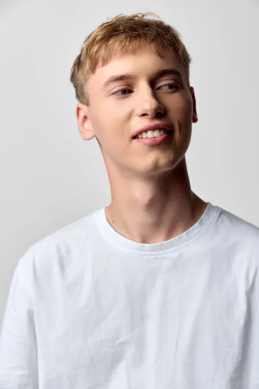 Young man wearing a white t shirt smiles softly against a neutral studio backdrop, creating a fresh, approachable mood for lifestyle, fashion and product promotion