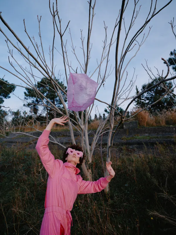 Outdoor scene with a person in a pink jacket throwing a pink scarf toward a bare tree, capturing motion and playful energy across a grassy field under a cloudy sky, emphasizing color, movement and